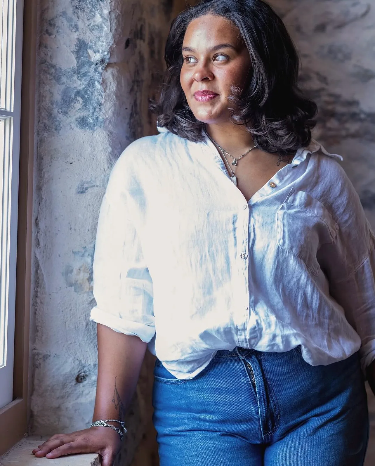 Natural-light portrait of a woman in a white linen shirt beside a stone wall window