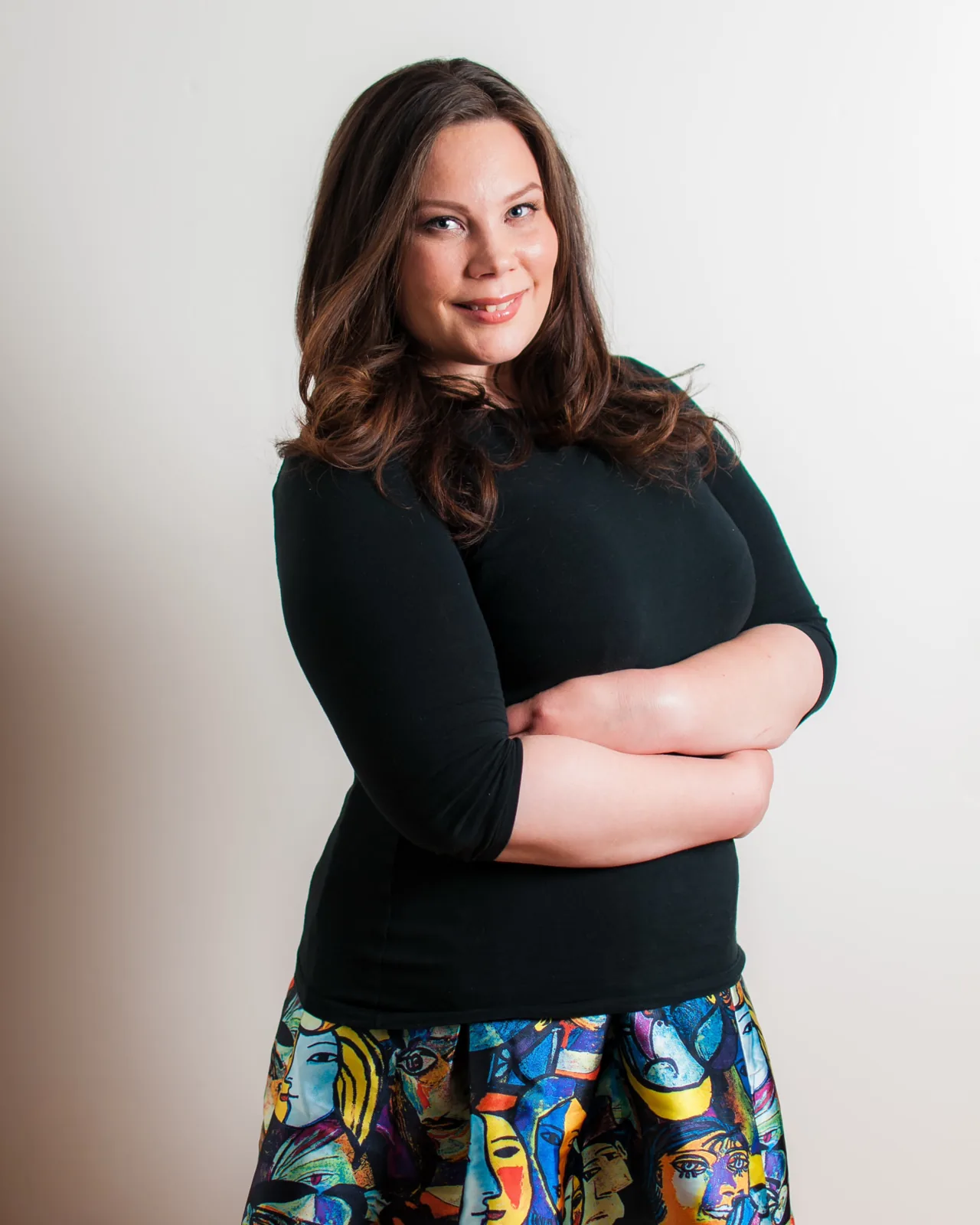 Studio headshot of a smiling woman in a black top and a colorful printed skirt against a light backdrop