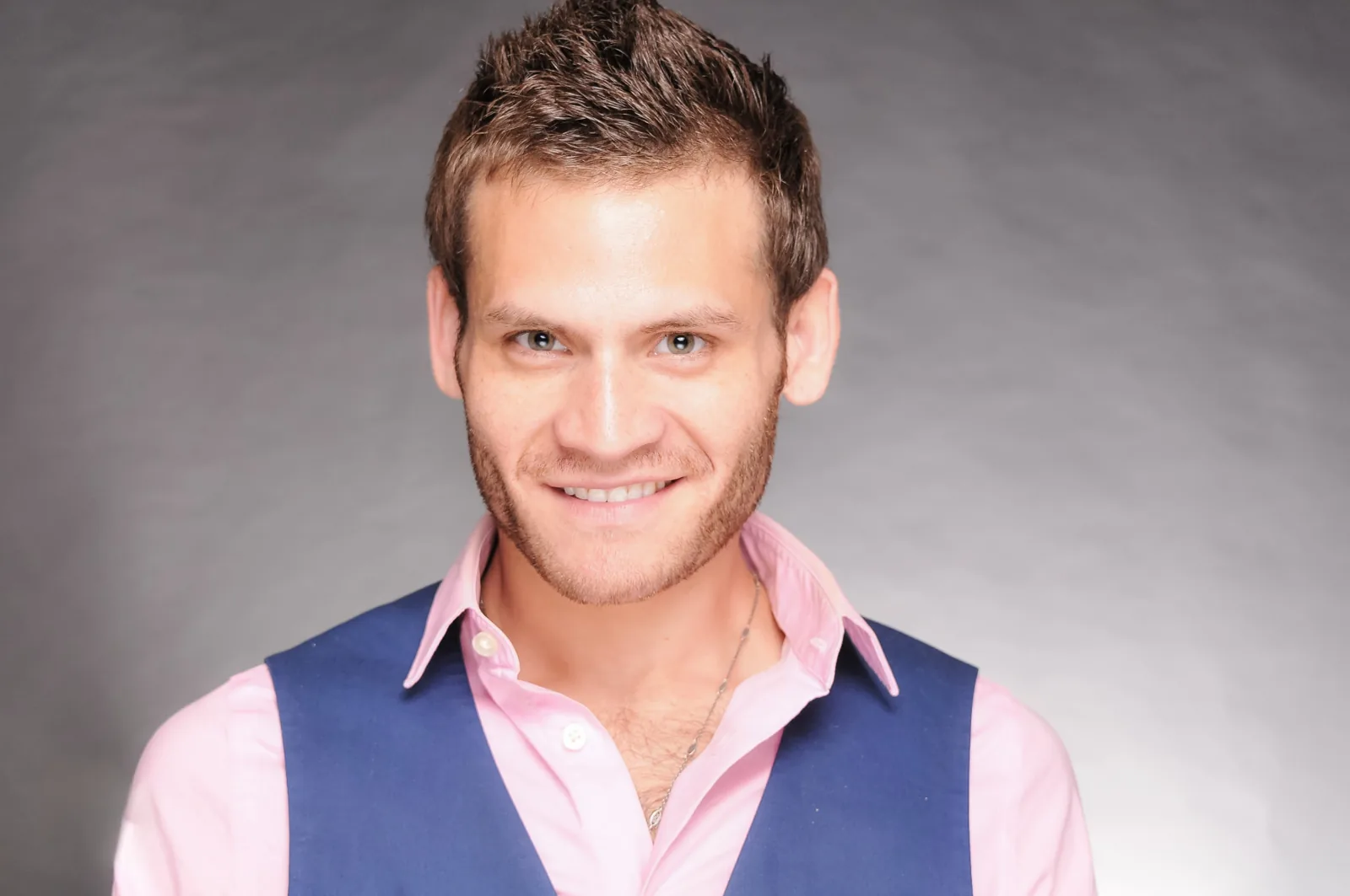 Studio headshot of a smiling man in a pink shirt and navy vest against a grey backdrop