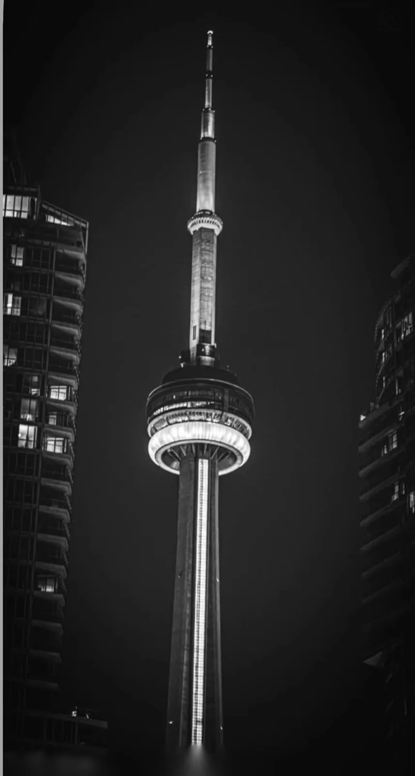 Black and white night photograph of a tall observation tower framed by two high-rise buildings