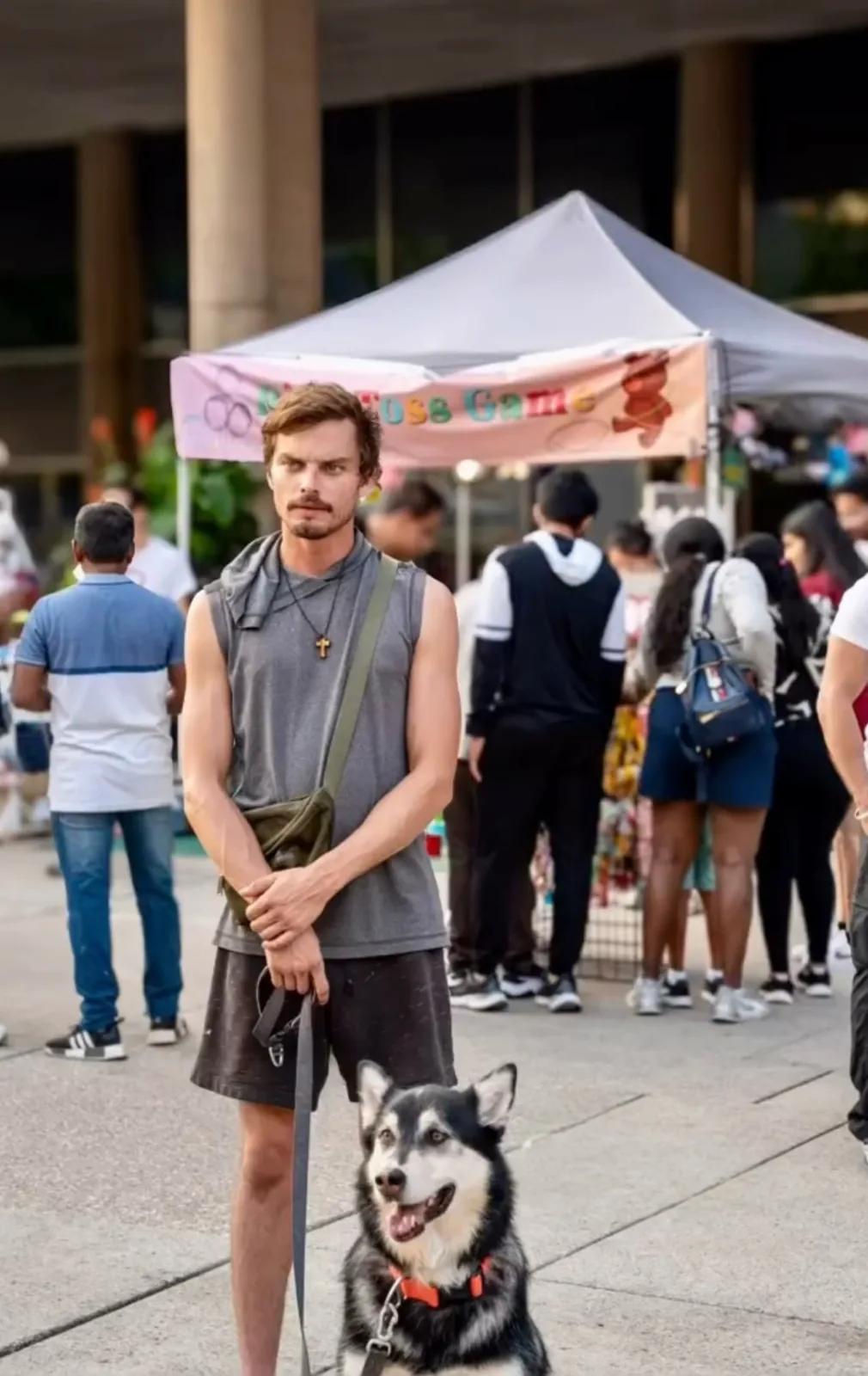 Man in a grey sleeveless top standing with a husky on a leash at an outdoor market