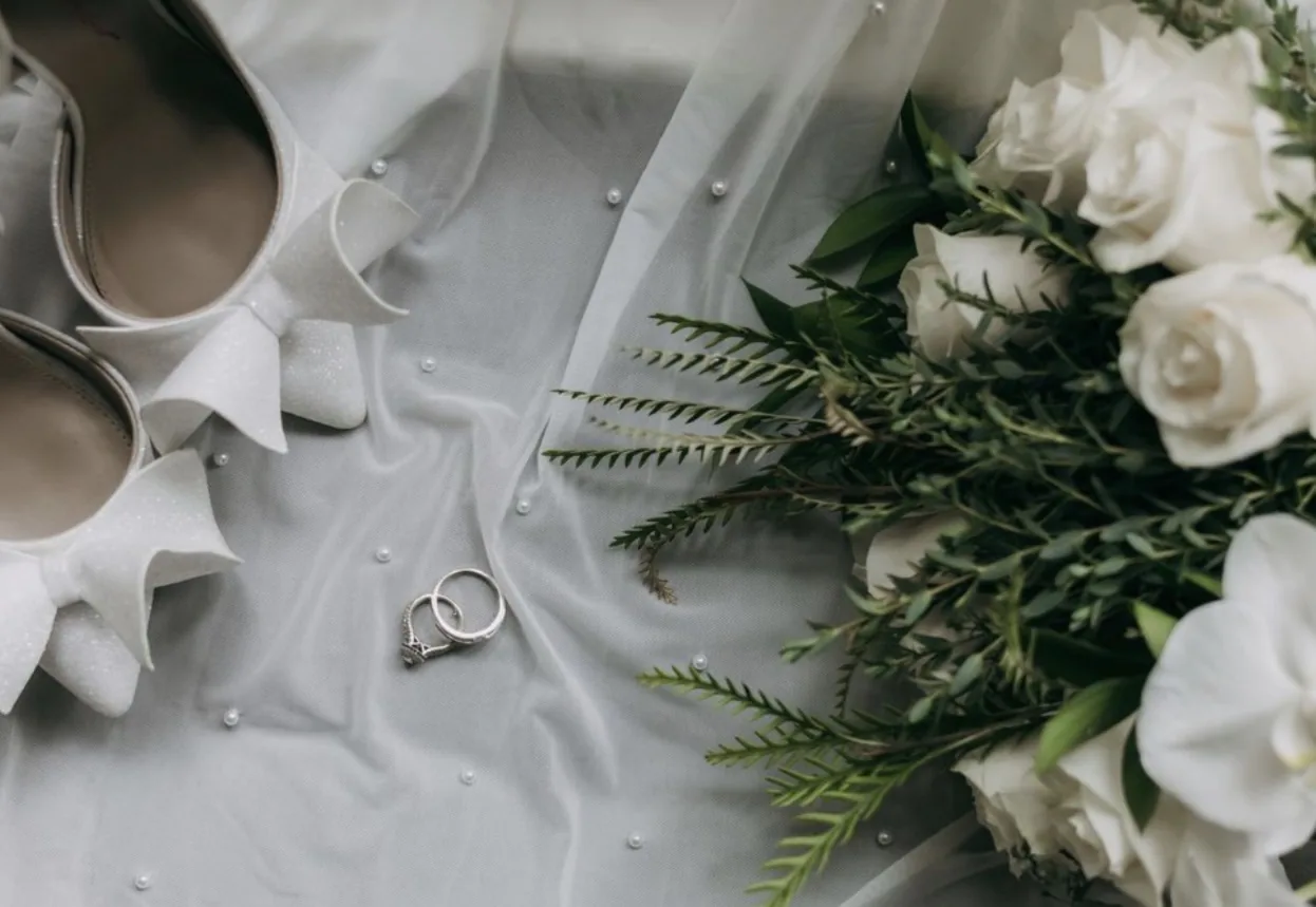 Wedding rings on a pearl veil beside white roses and bridal shoes