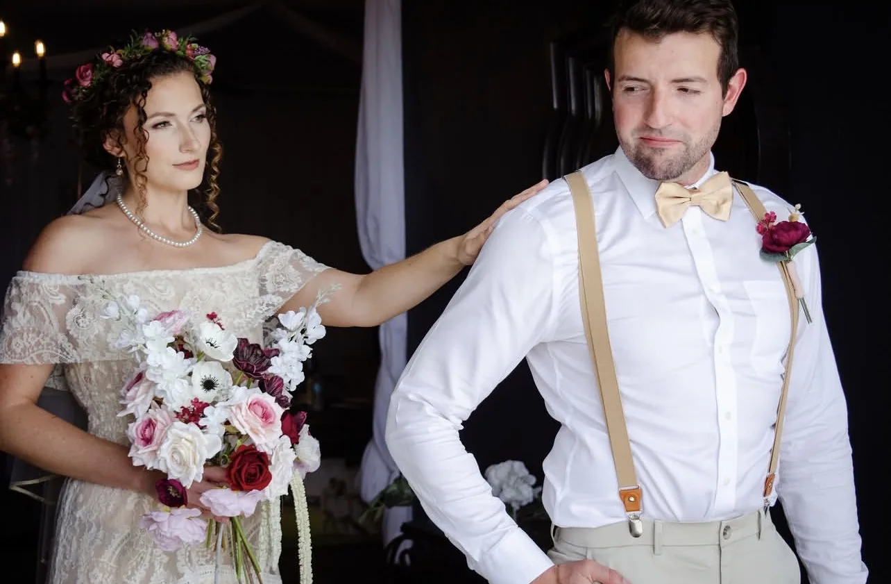 Wedding portrait of a bride in a lace gown with a floral crown resting her hand on the shoulder of a groom in suspenders and a bow tie