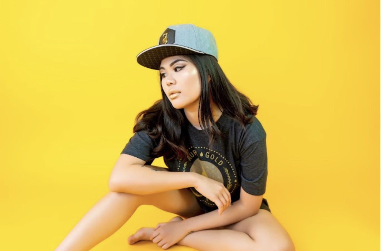 Seated studio portrait of a person in a cap and dark t-shirt looking to the side against a yellow backdrop