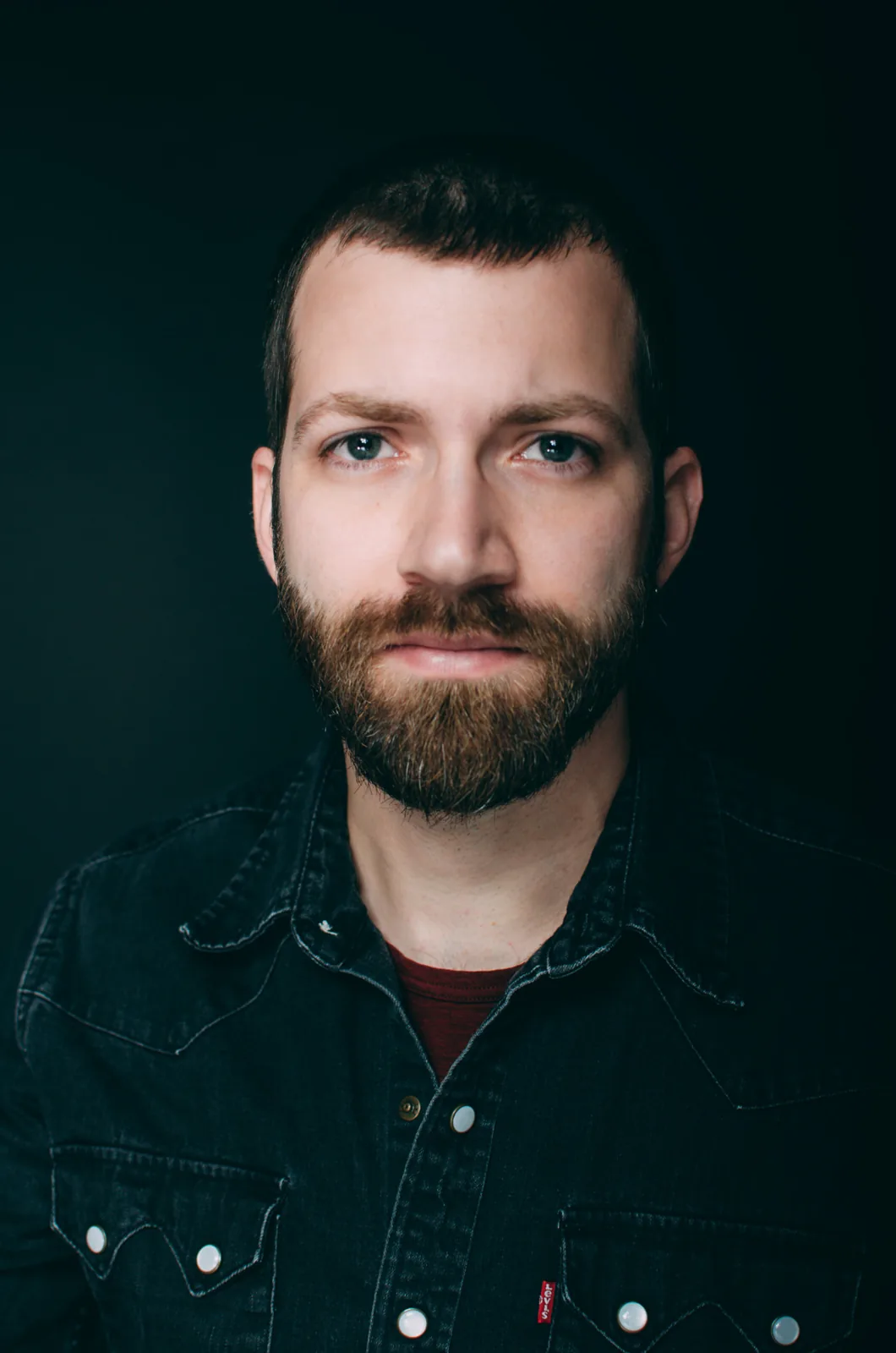 Studio portrait of a bearded man in a dark denim shirt against a black background