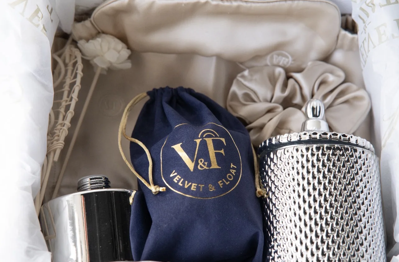 Close-up product photograph of a gift box with silver flasks, a satin scrunchie, an eye mask and a navy drawstring pouch