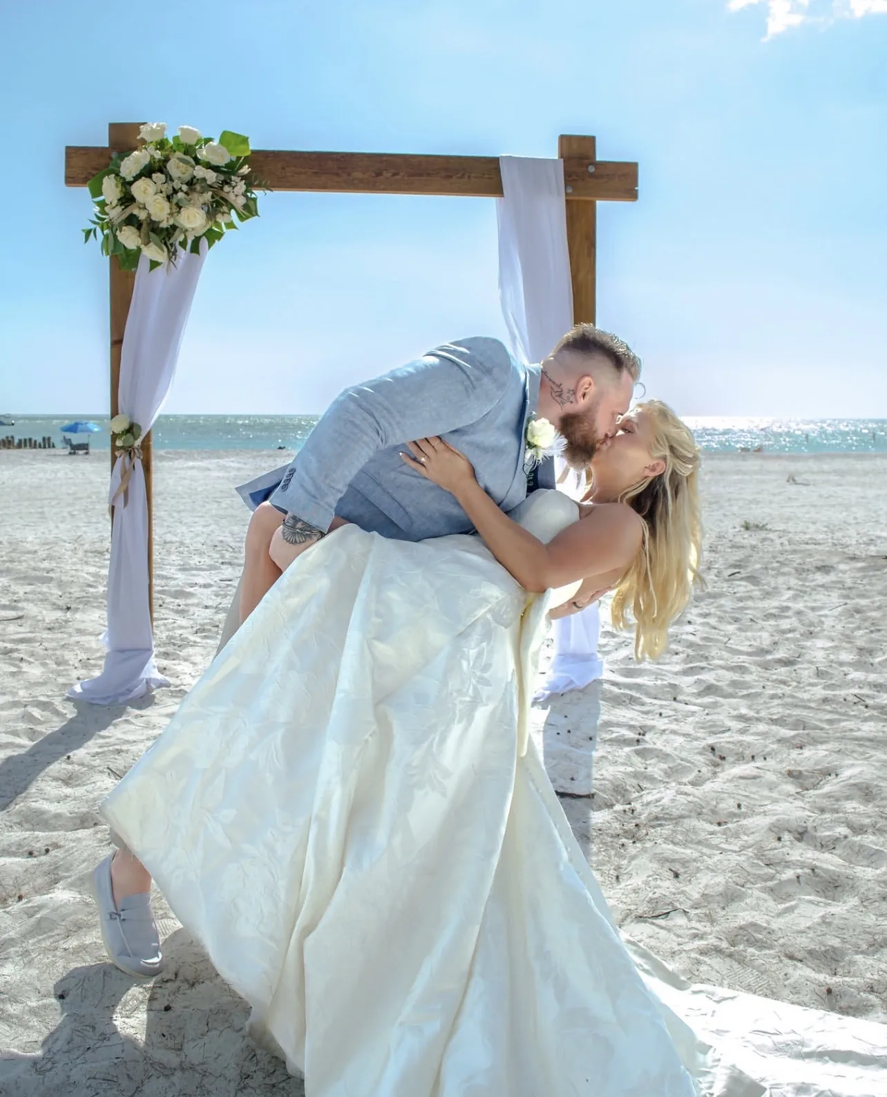 Beach wedding portrait of a groom dipping and kissing his bride beneath a floral arch by the ocean