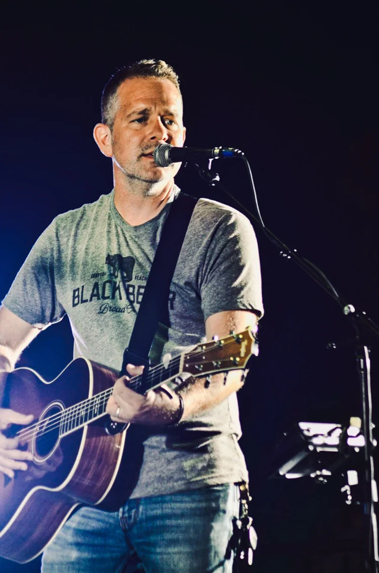 Musician singing into a microphone while playing acoustic guitar on stage
