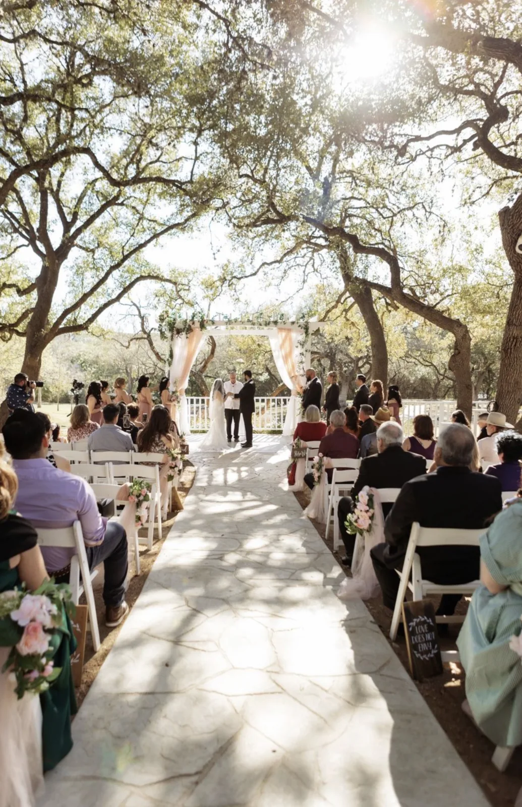Outdoor wedding ceremony beneath oak trees with guests seated along a stone aisle