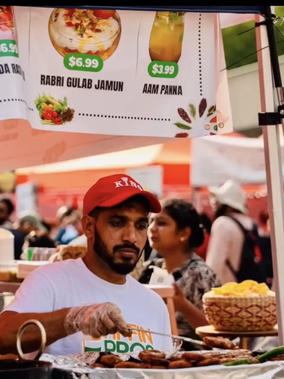 Food vendor in a red cap grilling skewers under a printed menu banner at a street food festival