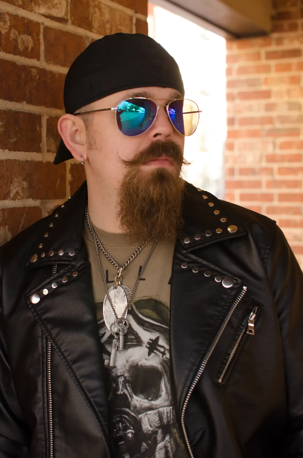 Editorial portrait of a man in a studded leather jacket and sunglasses beside a brick wall