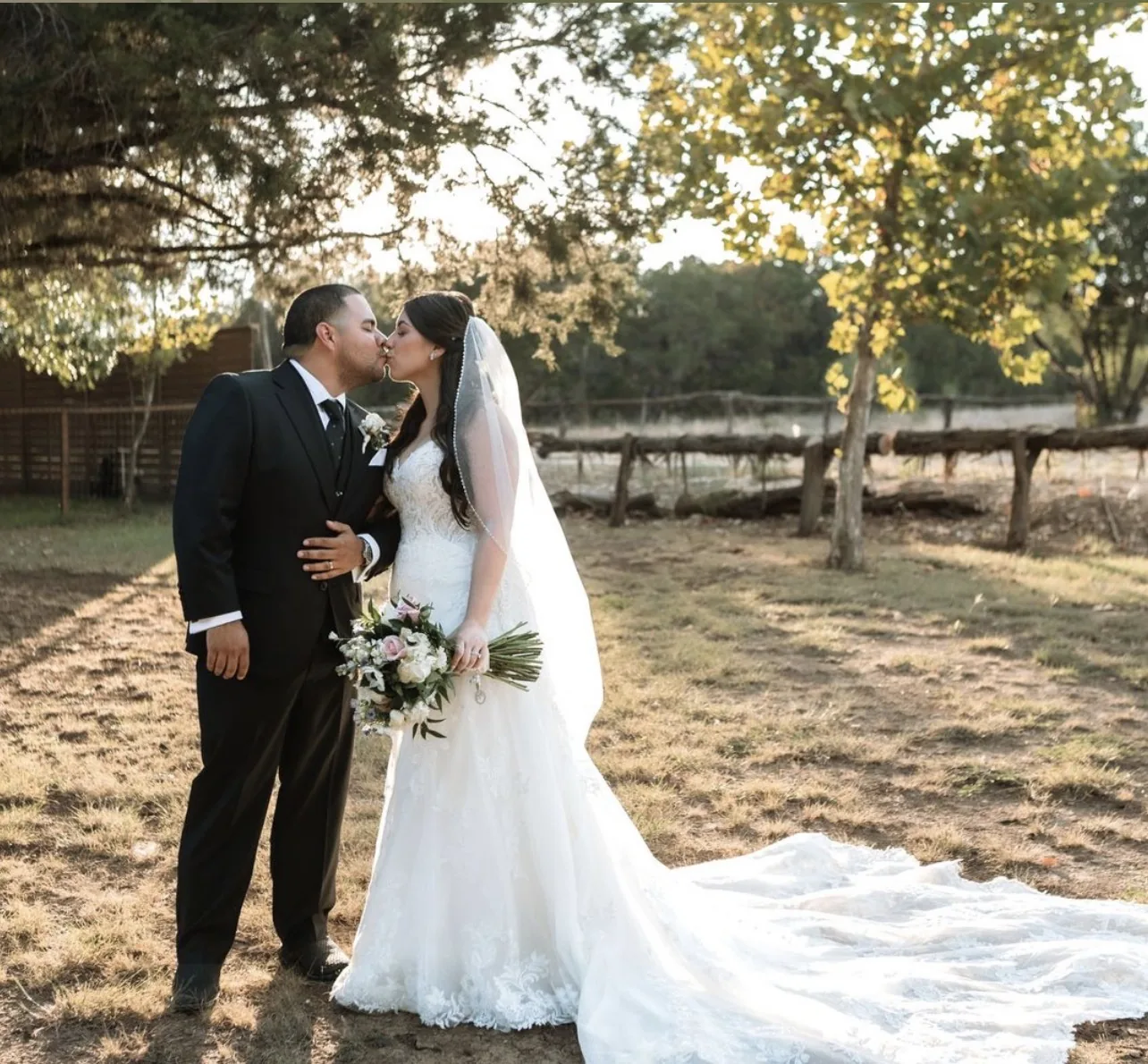 Bride and groom kissing beneath trees at golden hour with a long lace train