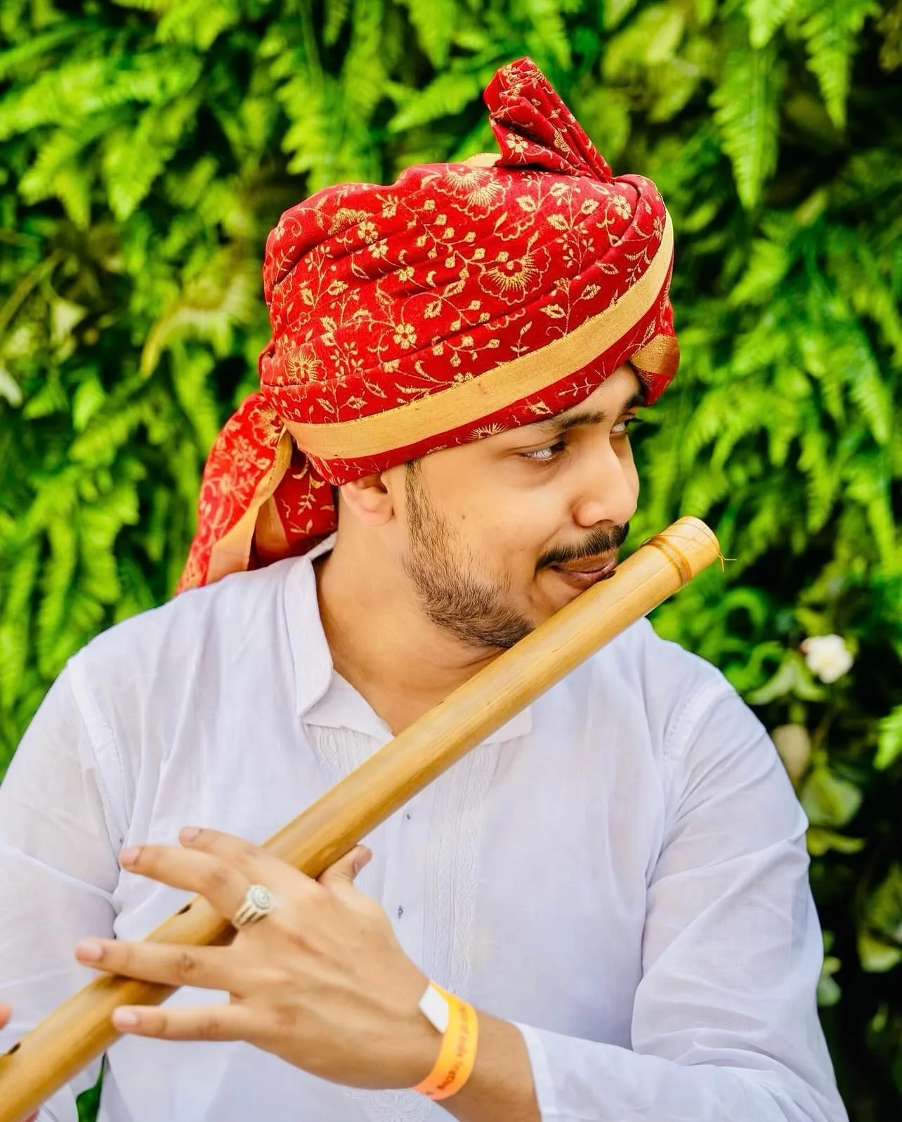 Man in a white kurta and red embroidered turban playing a bamboo flute in front of a green fern wall