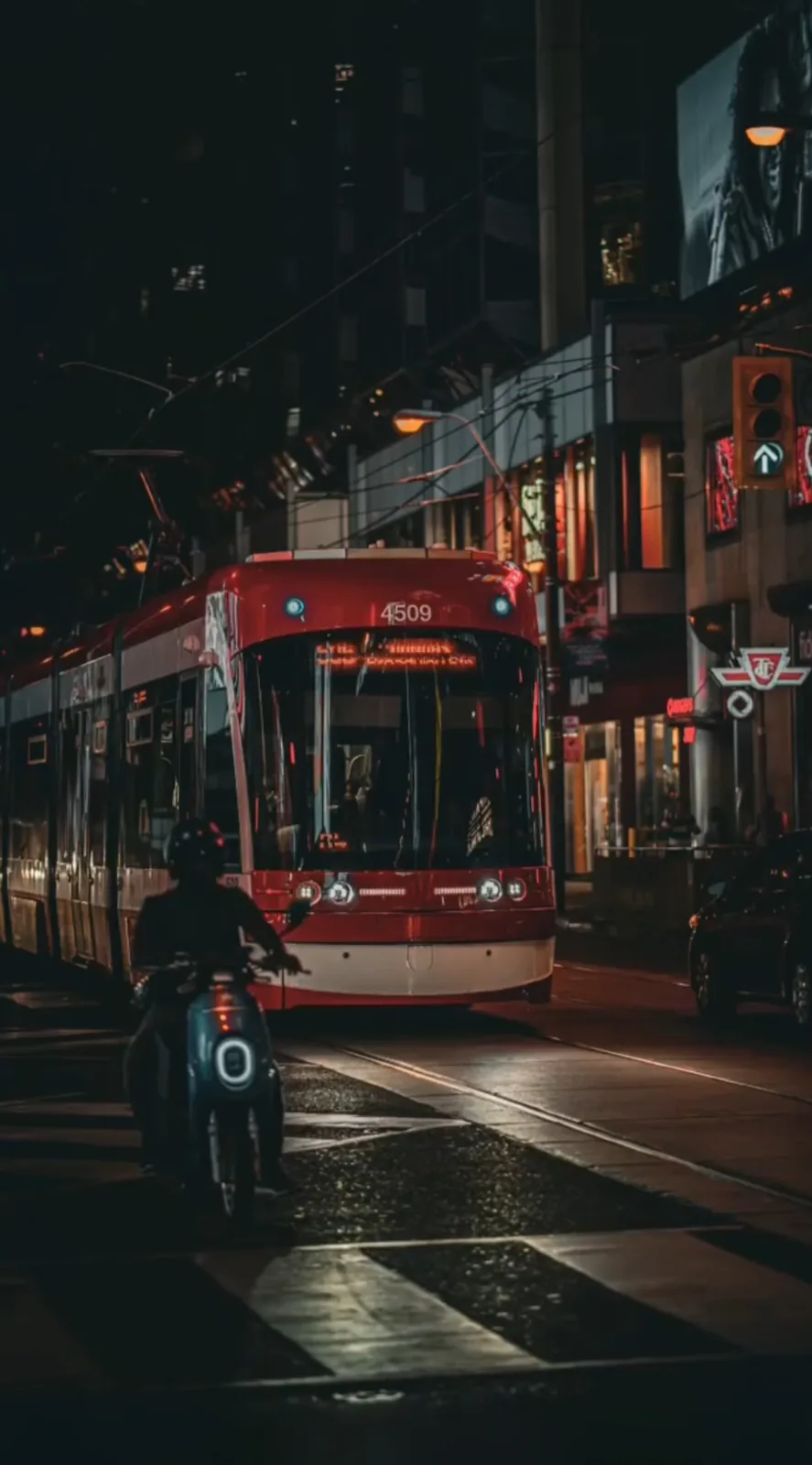 Red streetcar approaching a wet downtown intersection at night with a scooter rider waiting nearby