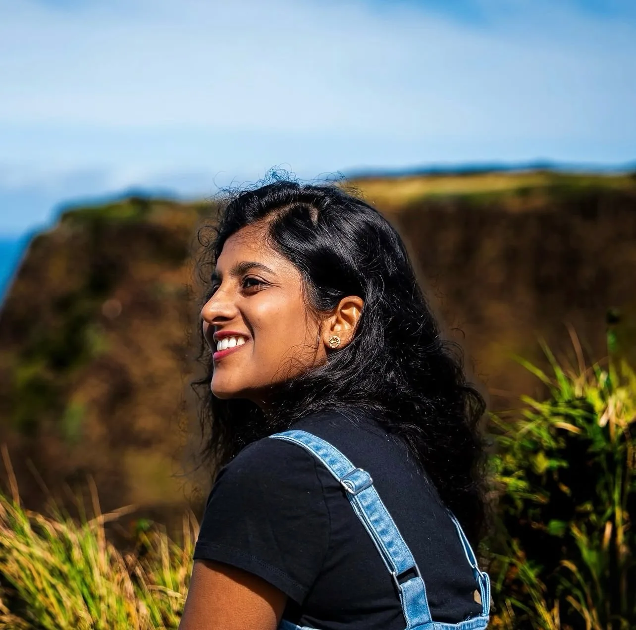 Smiling woman in a black tee and denim overalls looking back over her shoulder on a grassy clifftop