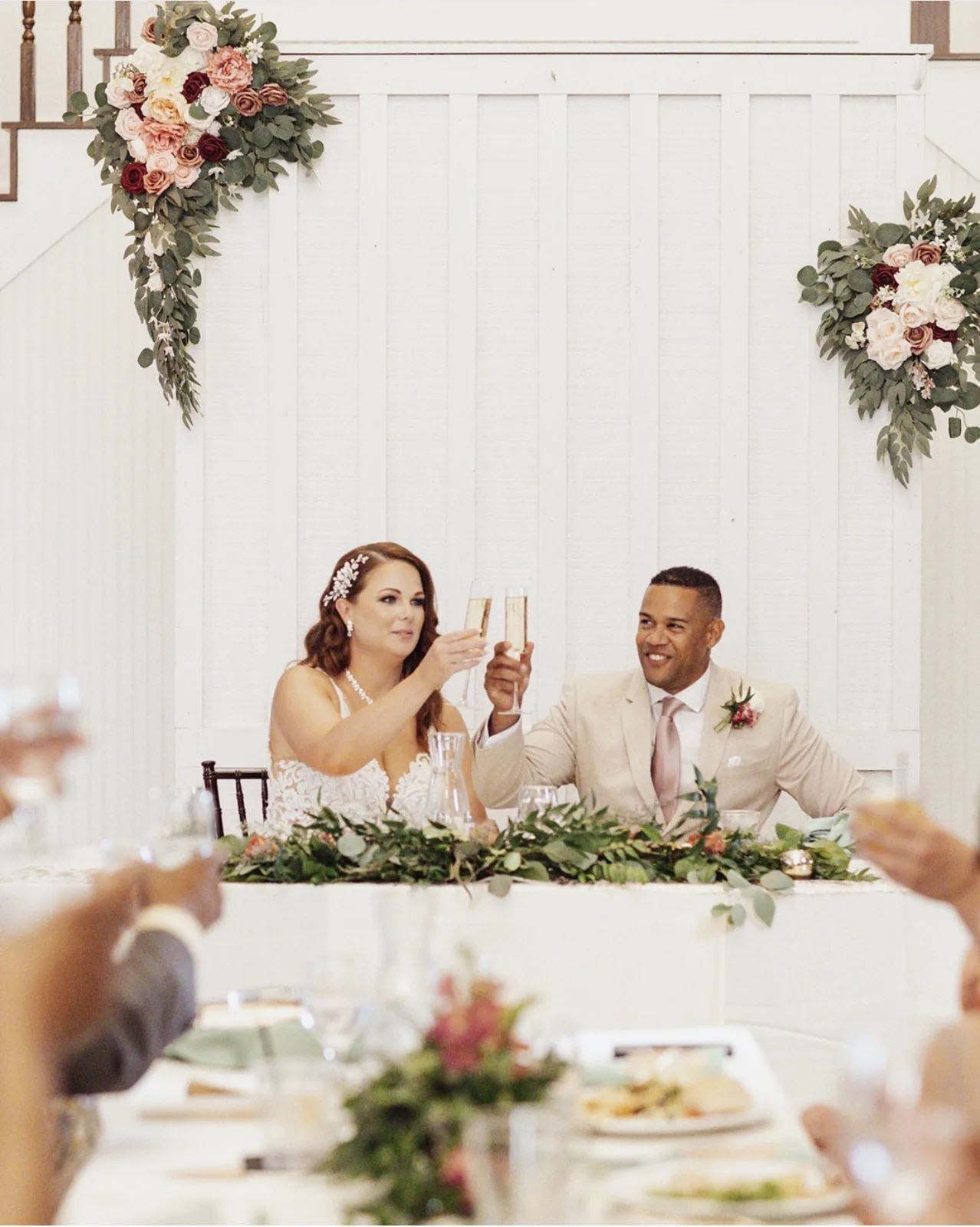 Newlyweds raising a champagne toast at a floral-draped reception head table