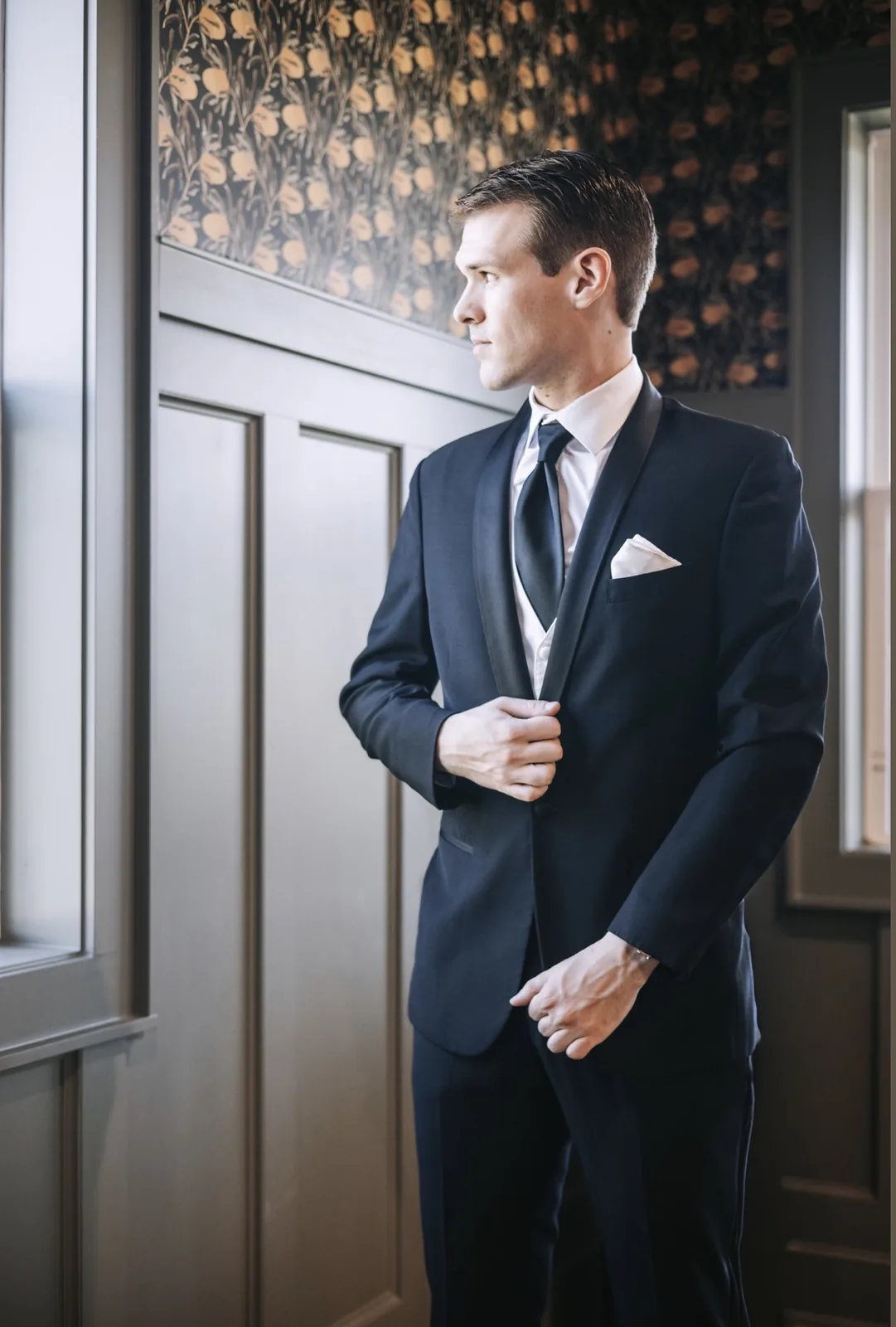 Window-light portrait of a groom adjusting his navy tuxedo jacket