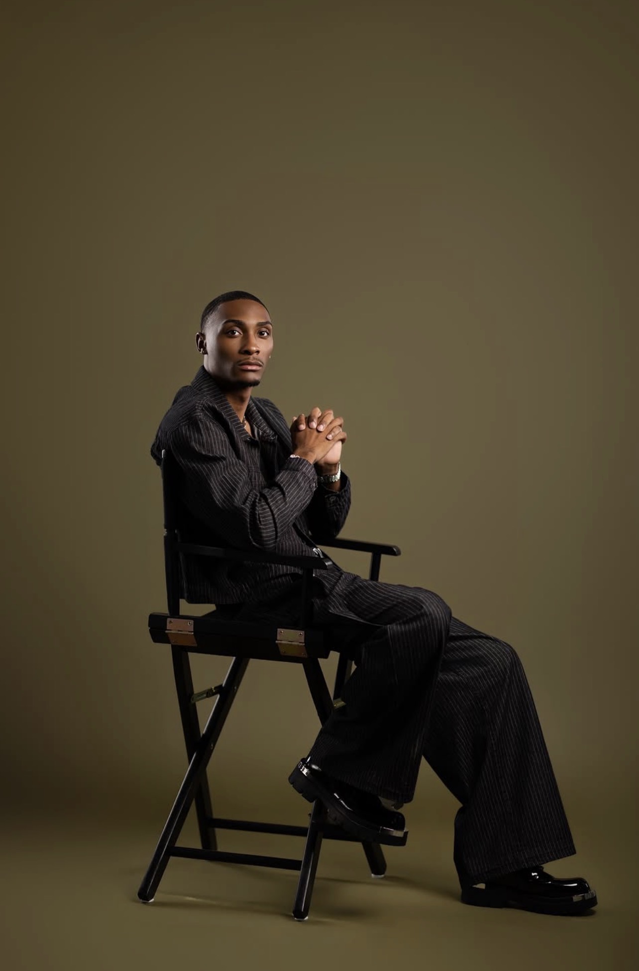 Editorial studio portrait of a man in a pinstripe suit seated in a director chair